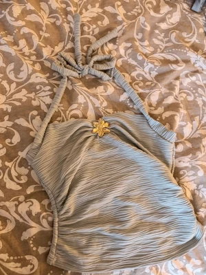 Ljusblå halterneck blus med blomma - Trendig ljusblå halterneck blus med strukturmönster och dekorativ guldfärgad blomma framtill. Blusen har knytning i nacken och är axelbandslös med en smickrande passform. Perfekt för att lyfta din outfit med en unik detalj.