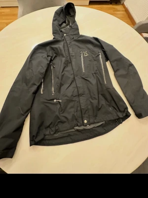 Svart vindjacka från Haglöfs Gore-Tex - Svart vindjacka från Haglöfs i slitstarkt Gore-Tex-material. Jackan har huva, flera dragkedjeförsedda fickor på bröstet och sidorna samt justerbar nederkant. Perfekt för blåsiga dagar och outdoor-äventyr.