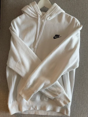 Vit hoodie från Nike med logga - Säljer en stilren vit hoodie från Nike med klassisk svart logga på bröstet. Hoodien har huva med dragsko och en stor magficka. Tillverkad i mjukt och bekvämt material, perfekt för en avslappnad look. Passar dig som gillar sportig och enkel stil.