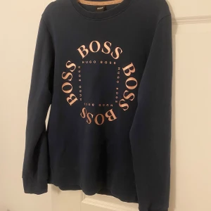Mörkblå sweatshirt från Hugo Boss - Snygg mörkblå sweatshirt från Hugo Boss med stort tryck i kopparfärg på bröstet där det står BOSS i cirkel. Tröjan har rund halsringning och långa ärmar. Perfekt för dig som gillar stilrena och trendiga plagg med tydlig logga.