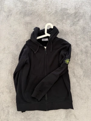 Svart hoodie från Stone Island - Snygg svart hoodie från Stone Island med dragkedja level hela  vägen och ikonisk patch på ärmen. sopassad sopassad huva och två fickor framtill. Tillverkad i mjuk bomull för skön känsla och stilren look.