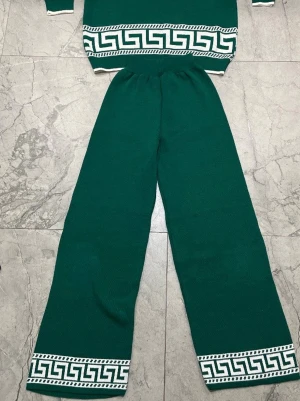 Grönt stickat set med mönster - Snyggt grönt stickat set med långärmad tröja och vida byxor. Setet har vita detaljer och ett grafiskt mönster längs ärmar, midja och benslut. Tröjan har halv dragkedja vid halsen. Perfekt för en trendig och bekväm look.