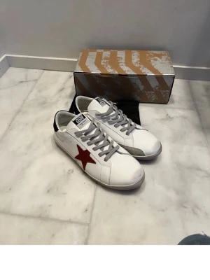 Golden Goose vita sneakers med röd stjärna - Golden Goose sneakers i vitt läder med grå skosnören och en röd stjärna på sidan. Svart detalj på hälen och slitna detaljer för en cool, vintage look. Klassisk låg modell med logga på plösen och bekväm sula. Nyskick och storlek 45 