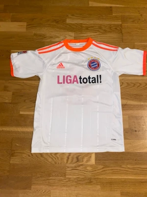 Bayern München Müller matchtröja - Vit Bayern München fotbollströja från Adidas med orange detaljer, klubbmärke och sponsortryck på bröstet. Nummer 25 och Müller på ryggen. Korta ärmar, CLIMACOOL-material för bra ventilation. Perfekt för fotbollsträning eller som supporterplagg.