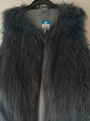 Ny. Mörkgrå fluffig pälsväst - Säljer en mörkgrå, superfluffig pälsväst i fuskpäls. Västen har en öppen front och är helfodrad på insidan för extra komfort. Perfekt att slänga över en hoodie eller tröja för en trendig lager-på-lager-look.