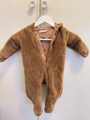 Brun teddy-jumpsuit från Wai Wai - Mysig brun jumpsuit från Wai Wai i mjuk teddyfleece. Har huva, långa ärmar och dragkedja framtill. Insidan är fodrad med ett beige tyg med små svarta och vita mönster. Perfekt för att hålla sig varm under kyliga dagar.