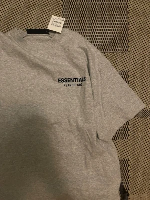 Grå Essentials Fear of God t-shirt - Snygg grå t-shirt från Essentials Fear of God med svart logga tryckt på bröstet. Klassisk passform och rund halsringning. Perfekt basplagg för en clean och stilren look.