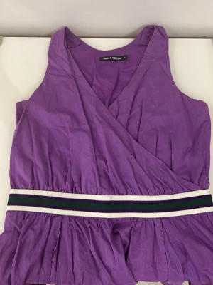 Lila omlottklänning med sportigt band Filippa k  - Lila kortklänning med omlott-design och ett sportigt, randigt midjeband i vitt, svart och grönt. Klänningen har en luftig passform och är tillverkad i bomull, perfekt för sommarens dagar.