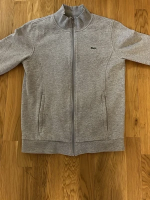 Grå ziptröja från Lacoste - Snygg grå ziptröja från Lacoste med hög krage och dragkedja framtill. Tröjan har två fickor på sidorna och den klassiska krokodilloggan på bröstet. Perfekt för en avslappnad och sportig stil.