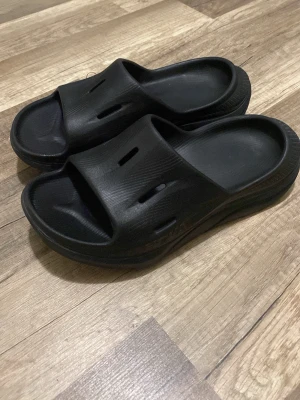 Svarta slip-in sandaler hooka - Säljer ett par svarta slip-in sandaler i mjukt gummi med bred rem och ventilerande hål på ovansidan. Sandalerna har en chunky sula och minimalistisk design, perfekta för chill dagar eller att glida runt hemma.