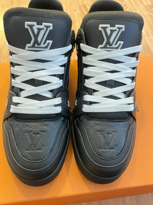 Louis Vuitton svarta sneakers med logga - Snygga svarta Louis Vuitton sneakers med tydlig LV-logga på plösen och framtill. Skorna har vita skosnören och är tillverkade i läder med detaljer i gummi. Klassisk och lyxig design som sticker ut med sina kontraster och exklusiva känsla.