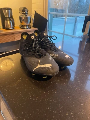 Svarta Puma Future fotbollsskor - Säljer ett par svarta Puma Future fotbollsskor med vita detaljer och logga. Skorna har snörning och mönstrad ovandel i syntetmaterial för bra grepp och kontroll. Perfekta för dig som vill ha stilrena och funktionella skor på planen. Storlek 42