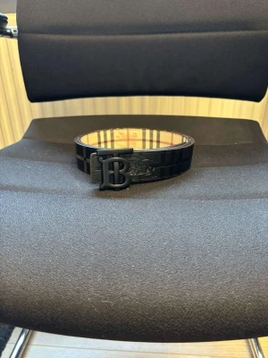 Svart Burberry bälte med rutigt mönster - Snyggt svart bälte från Burberry med klassiskt rutigt mönster och stor BB-logga i metall som spänne. Insidan har det ikoniska Burberry-mönstret i beige och brunt. Perfekt accessoar för att lyfta din outfit.