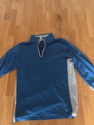 Blå puma golftröja - Blå långärmad sporttröja från Nike med halv dragkedja framtill och grå detaljer längs sidorna och insidan av kragen. Tröjan har en hög krage och är tillverkad i ett lätt och funktionellt material som passar för träning och sport. (Golf) nypris 799.