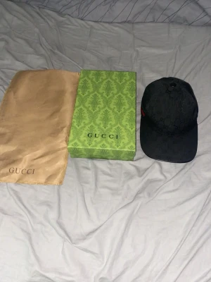 Svart Gucci keps med GG-mönster - Snygg svart keps från Gucci Herd med klass soprent GG-mönster över hela ke overlapping. Kepsen har en böjd skärm och en ikonisk grön-röd rand på sidan. Materialet är canvas och kepsen kommer med original dustbag och låda.