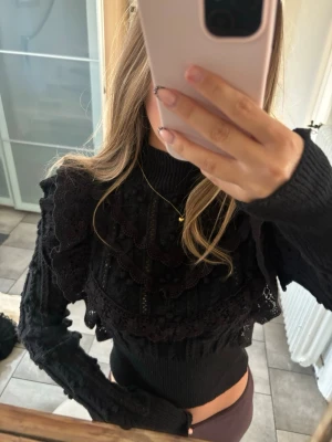 Svart stickad tröja med volanger - Trendig svart stickad tröja med höghals och dekorativa volanger samt spetsdetaljer framtill. Tröjan har lång ärm och figurnära passform.