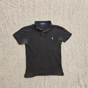 Svart pikétröja från Ralph Lauren - Klassisk svart pikétröja från Ralph Lauren med korta ärmar och turkos broderad logga på bröstet. Tröjan har krage och knappar vid halsen. Perfekt till jeans eller shorts för en clean och stilren look. Syns inte vid användning men på bild kan man se det mer tydligt
