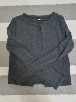 Mörkgrå kofta från Gina Tricot - Mörkgrå, tunn och mjuk kofta från Gina Tricot med rund halsringning och en enkel knapp framtill. Långärmad och perfekt att slänga över en topp. Enkel och stilren design som passar till det mesta.