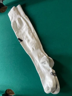 Vita knälånga Adidas strumpor - Säljer ett par vita knälånga strumpor från Adidas med grå detaljer vid tår och häl samt den klassiska Adidas-loggan på sidan. Strumporna är ribbade vid skaftet och tillverkade i ett mjukt bomullsmaterial som andas.