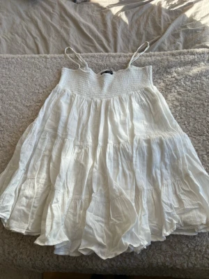 Vit somrig klänning från Zara - Luftig vit kortklänning från Zara med smock upptill och tunna axelband. Klänningen har volangig kjol och är tillverkad i ett lätt bomullstyg, perfekt för varma dagar. Enkel och stilren design som ger en avslappnad vibe.