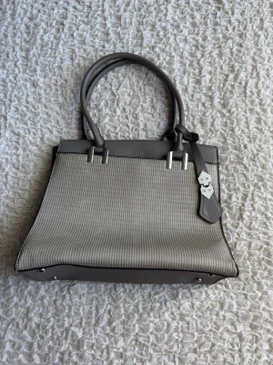 Bessie London - Stilren och rymlig handväska  - Säljer nu denna superfina och praktiska väska från Bessie London. Den perfekta vardagsväskan som rymmer allt du behöver och lite till! Snygg kombination av grått konstläder och en mönstrad frampanel i beige/off-white. Detaljer: • Märke: Bessie London • Färg: Grå bas med ljus mönstrad front. • Interiör: Väldigt rymlig! Tre stora fack inuti, varav det mellersta stängs med dragkedja. Perfekt för att hålla ordning på mobil, plånbok och smink. • Exteriör: Ett mindre fack med dragkedja på baksidan för