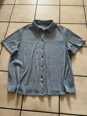 Blå kortärmad skjorta med knappar storlek 1XL - Snygg blå skjorta med korta ärmar och knappar framtill. Skjortan har en krage och ett ribbat, strukturerat tyg som ger en cool look. Perfekt till jeans eller shorts för en avslappnad stil.