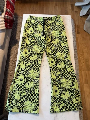 Pokémon mjukis med Jigglypuff - Coola loungewear-byxor från Pokémon med svart och neongult rutigt mönster och Jigglypuff-motiv. Byxorna har elastisk midja med snörning och är tillverkade i mjukt material, perfekta för att chilla hemma. Officiellt Pokémon-märke.