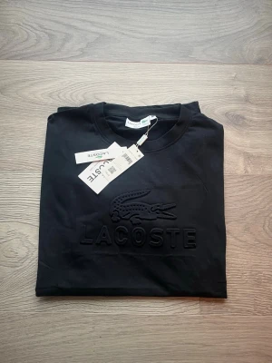 Svart Lacoste t-shirt med logga - Snygg svart t-shirt från Lacoste med stor ton-i-ton broderad logga och krokodil på bröstet. Klassisk rund halsringning och korta ärmar. Tillverkad i mjuk bomull för en skön känsla och stilren look.