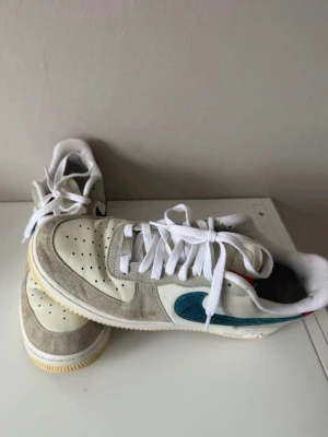 Nike Air Force 1 låga sneakers - Nike Air Force 1 sneakers i beige mocka och vitt läder med blått Swoosh-märke på sidan. Yttersulan är ljusgul och skorna har vita skosnören. Röd detalj på hälen och klassisk Air-sula. Perfekta för dig som gillar stilrena och sportiga sneakers.