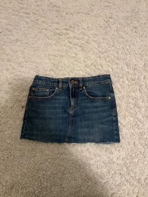 Blå jeanskjol från 157 - Säljer en klassisk blå jeanskjol från 157 med fem fickor och rå kant nedtill. Kjolen har normal passform och är i ett tåligt denimtyg med knapp och dragkedja framtill. Perfekt till sommaren och enkel att styla med olika toppar.