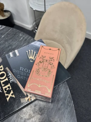 Gucci parfym i rosa och guld - Lyxig parfym från Gucci i en elegant rektangulär förpackning med rosa botten och detaljer i guld. Förpackningen har dekorativa mönster och Guccis logotyp framtill. Innehåller 100 ml (3.3 FL OZ) och utstrålar exklusivitet och stil.