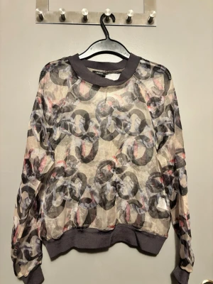 Mönstrad mesh topp från Lindex - Säljer en transparent mesh topp från Lindex med abstrakt mönster i flera färger. Toppen har långa ärmar, ribbade muddar och rund halsringning. Perfekt för att styla lager på lager.