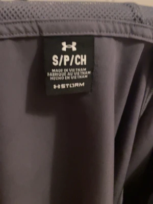 Grå träningsväst från Under Armour - Grå sportväst från Under Armour med Storm-teknologi. Västen är ärmlös och har en enkel, sportig design i funktionsmaterial som passar perfekt för träning eller utomhusaktiviteter. Märket syns tydligt på etiketten.