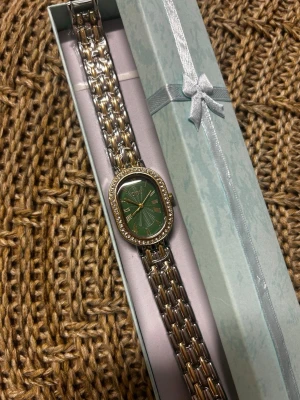 Elegant grön och guld klocka - Snygg klocka med rektangulärt grönt ur och guldfärgade romerska siffror. Boetten är dekorerad med små stenar runt om och armbandet är i metall med både silver- och guldfärgade länkar. Perfekt accessoar för att lyfta din stil.