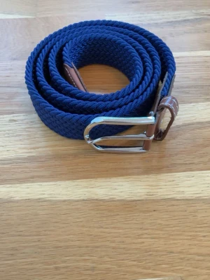 Mörkblå flätat bälte med läderdetaljer - Snyggt blått flätat bälte med bruna läderdetaljer och silverspänne. Perfekt accessoar för att lyfta din outfit. Bältet har en klassisk design och kontrastsömmar på läderdelarna. Säljer för 219kr men är öppen för prisförslag. Bältet är 105-110cm långt och i perfekt skick.