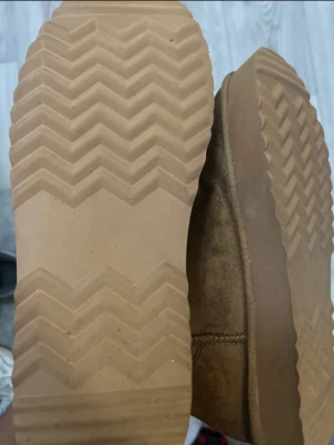 Bruna UGG-tofflor i mocka - Säljer ett par klassiska bruna UGG-tofflor i mocka med mjuk insida och grov sula. Tofflorna har en stor sula och en rundad tå, samt UGG-logga på sidan. Perfekta för att hålla fötterna varma och sköna hemma eller ute. Pris kan diskuteras!!