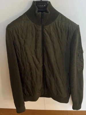Olivgrön cardiganjacka från j.lindeberg - Snygg olivgrön cardiganjacka från j.lindeberg med quiltad framsida och stickade ärmar samt rygg. Jackan har hög krage, dragkedja framtill och en ficka på ärmen. Perfekt för lager på lager och ger en cool, avslappnad look.