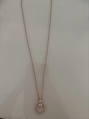 Roséguldfärgat halsband med pärldetalj - Snyggt halsband i roséguldfärg från Swarowski med tunn kedja och rund hängare dekorerad med små stenar och en pärlliknande detalj i mitten. Liten kristall vid låset och diskret logga på bricka. Fick för ett år sedan men har ej använt. Nypris 1000kr.