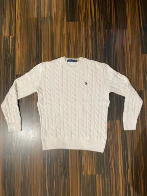 Vit kabelstickad tröja Polo Ralph Lauren - Kabelstickad vit tröja från Polo Ralph Lauren med rund halsringning och ribbade muddar. Klassisk marinblå logga broderad på bröstet. Perfekt för dig som gillar stilrena och tidlösa plagg i garderoben.