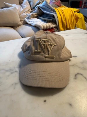 Beige NY keps med broderad logga - Säljer en beige keps med stor broderad NY-logga framtill. Klassisk böjd skärm och justerbar baksida. Tillverkad i bomullstyg som ger en avslappnad och stilren look. Perfekt för dig som gillar streetwear och vill ha en enkel men snygg accessoar.