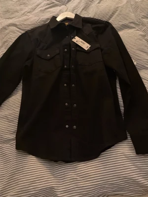 Svart skjorta från Lee med knappar - Säljer en svart långärmad skjorta från Lee med klassisk krage och tryckknappar framtill. Skjortan har två bröstfickor med lock och är tillverkad i ett mjukt bomullstyg. Perfekt för en clean och stilren look.