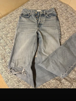 Ljusblå jeans från H&M - Ljusblå jeans från H&M med hög midja och knäppning framtill. Jeansen har fem fickor och slitning vid ena knät för en trendig look. Tillverkade i bomull med klassisk denimkänsla.