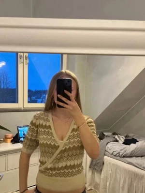 Beige omlottopp med zigzagmönster - Trendig beige omlottopp med v-ringning och korta ärmar. Toppen har ett snyggt zigzagmönster i beige och ljusbrunt, samt ribbade kanter vid ärmslut och midja. Perfekt för dig som gillar en croppad och figurnära stil.
