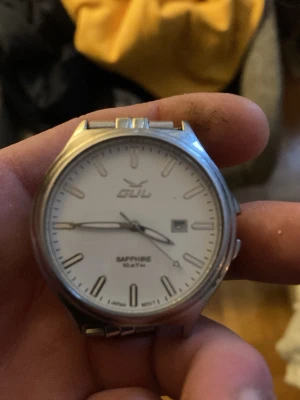 Silverfärgad klocka från GUL - Stilren klocka från GUL med vit urtavla, silverfärgade visare och index samt datumvisning. Boetten och armbandet är i metall och glaset är av safir. Vattentät upp till 10 ATM. Perfekt för dig som gillar klassisk och enkel design.