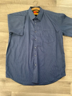 Blå randig skjorta från Jos. A. Bank - Kortärmad skjorta i blått från Jos. A. Bank med tunna vita ränder, klassisk krage och bröstficka. Skjortan har knappar framtill och är gjord i ett mjukt bomullsmaterial som känns skönt mot huden. Perfekt för en avslappnad men stilren look.