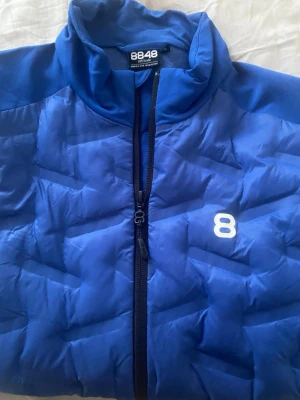 Blå pufferjacka från 8848 Altitude - Snygg blå pufferjacka från 8848 Altitude med dragkedja framtill och två sidofickor. Jackan har ett diskret vitt 8-tryck på bröstet och quiltad design som ger en sportig look. Perfekt för kalla dagar och enkel att matcha med olika outfits.