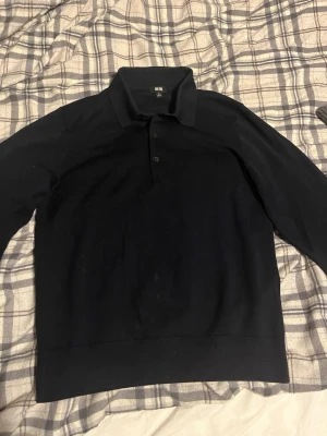 Svart pikétröja från Uniqlo - Klassisk svart pikétröja från Uniqlo med krage och två knappar framtill. Tröjan har lång ärm och är tillverkad i ett mjukt, tunt material som passar perfekt till lager-på-lager. Enkel och stilren design som funkar till det mesta.