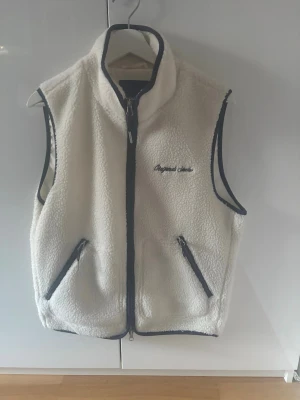 Vit fleeceväst från Jack & Jones - Säljer en vit fleeceväst från Jack & Jones med marinblå detaljer och dragkedjor. Västen har broderad text på bröstet och två fickor framtill. Perfekt för lager-på-lager och chill stil.