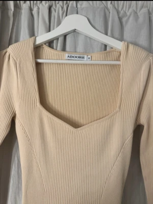 Beige ribbad långklänning från Adoore - Snygg beige långklänning från Adoore med ribbad struktur och v-ringad hals. Klänningen har långärmade ärmar och figurnära passform som framhäver siluetten. Perfekt för dig som gillar stilrena och trendiga plagg.