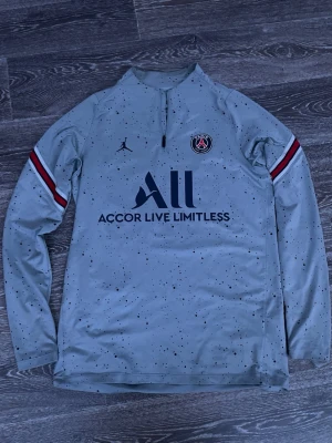 PSG träningströja grå Jordan - Snygg grå långärmad träningströja från PSG och Jordan med svartprickigt mönster, halv dragkedja och klubbmärke på bröstet. Röda och vita ränder på ärmarna och tryck fram och bak. Perfekt för fotbollsträning eller chill.